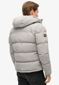 Chaqueta acolchada de color gris claro con capucha, que presenta un diseño acolchado, detalles de logotipo en la manga y cierre con cremallera. Jeans negros visibles.