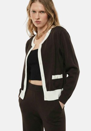 Donna con cardigan marrone con bordatura bianca, top corto nero, pantaloni marroni abbinati, collana a catena dorata, mani in tasca, su sfondo bianco.