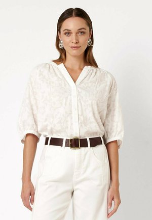 Frau mit braunen Haaren trägt eine bestickte weiße Bluse mit Puffärmeln, hoch taillierte weiße Hose, einen braunen Gürtel und Perlencluster-Ohrringe.