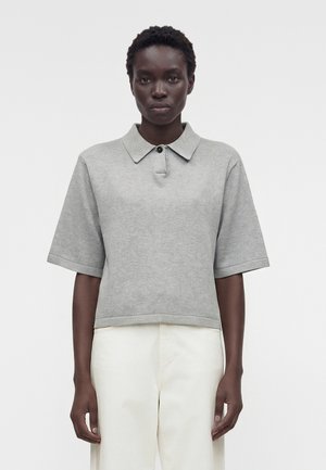 Γυναίκα φοράει ανοιχτογκρι κοντομάνικο πόλο πουλόβερ και ψηλόμεσο παντελόνι εκρού με απλό ανοιχτό φόντο.