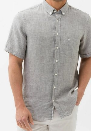 Homme portant une chemise à manches courtes gris clair à boutons avec un motif à carreaux subtil, associée à un pantalon blanc, une main dans la poche.