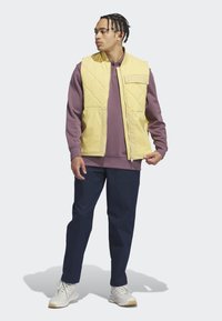 Žltá prešívaná vesta s prednými vreckami, oblečená cez fialovú mikinu, spárovaná s námorníckymi nohavicami a bielymi teniskami. Viditeľné textúrované materiály.