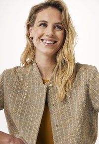 Jasnobrązowa kurtka o teksturowanej strukturze z dopasowanym krojem, złotymi akcentami i okrągłym dekoltem, noszona na musztardowej bluzce.