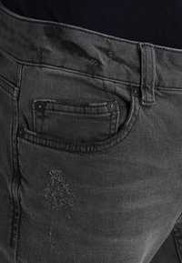 Svarta jeans i denim med en texturerad yta, utrustade med en framficka med metallbeslag och ett diskret slitet mönster på låret.