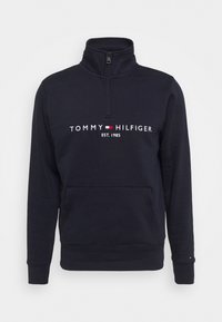 Felpa blu navy con colletto alto, chiusura con zip frontale e tasca anteriore. Presenta il logo "TOMMY HILFIGER" in bianco, rosso e blu. Tessuto morbido.