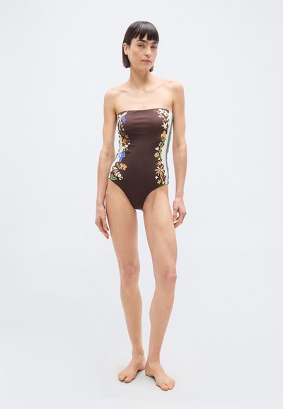 Γυναίκα που φοράει ένα καφέ ολόσωμο μαγιό χωρίς τιράντες με πολύχρωμα πλαϊνά σχέδια, όρθια ξυπόλυτη μπροστά σε απλό φωτεινό φόντο.