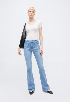 Γυναίκα με κοντά ξανθά μαλλιά που φοράει λευκό εφαρμοστό μπλουζάκι, ανοιχτόχρωμο τζιν ψηλόμεσο, μαύρα μυτερά παπούτσια και κρατάει μαύρη τσάντα ώμου.