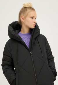 Młoda kobieta o blond włosach związanych w koka, ubrana w czarną kurtkę puchową na fioletowym dzianinowym swetrze, patrząca w prawo na tle jednolitego tła.