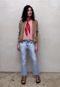 Femme aux cheveux bouclés portant une chemise rayée rouge, une écharpe rouge, une veste beige, un jean bleu clair et des sandales beiges, debout contre un mur uni.