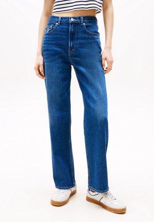 Personne portant un jean bleu taille haute, coupe droite, des baskets blanches à semelles marron et un top rayé sans manches court.