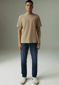 Tricou bej cu guler rotund, blugi drepți de culoare albastru denim și adidași albi cu talpă maro. Design minimalist și textură din bumbac.