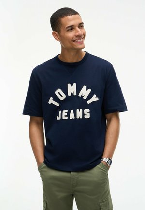Mand smilende, kiggende mod højre iført en mørkeblå "Tommy Jeans" T-shirt, olivengrønne cargobukser og sølvfarvet armbåndsur med hænderne i lommerne.