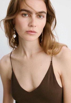 Joven mujer con cabello castaño claro, con pendientes geométricos dorados y un top marrón sin mangas, mirando directamente al frente con una expresión neutral.