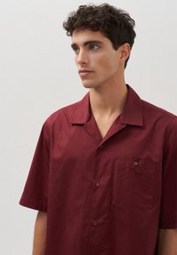 Camicia corta a maniche corte color mogano realizzata in tessuto leggero, con chiusura a bottoni frontale, tasca sul lato sinistro del petto e colletto con punte.