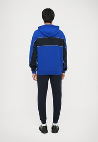 Hoodie u plavo-crnim bojama s kapuljačom, rebrastim manžetama i opuštenim krojem, uparen s uskim crnim hlačama i crnim tenisicama.