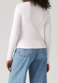 Frau, die ein enganliegendes weißes, geripptes Langarmoberteil und weite, hellblaue Jeans trägt, von hinten vor einem schlichten Hintergrund gezeigt.