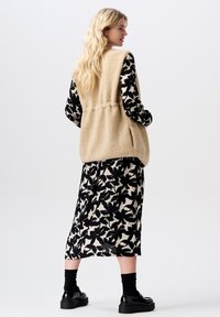 Beige Fleece-Weste mit Seitentaschen, über einem schwarz-creamfarbenen floral gemusterten Kleid getragen, kombiniert mit schwarzen Socken und Plateauschuhen.
