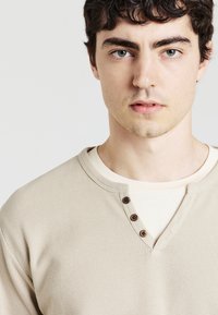 Maglietta henley beige a maniche lunghe in tessuto a costine, con tre bottoni marroni a livello del collo e un sottogiacca chiaro a contrasto.