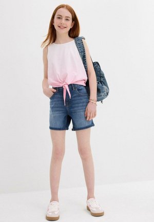 Haut sans manches rose et blanc noué à la taille, associé à un short en jean bleu. Accessoirisé avec un sac à dos en denim et des baskets légères.