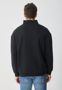 Sweatshirt preto com gola alta, punhos canelados e ajuste relaxado. Apresenta uma textura lisa e um design minimalista. Usado com jeans azuis.