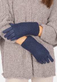 Marineblaue gerippte Handschuhe mit gestrickter Textur und Fingeröffnungen, die über einem hellgrauen flauschigen Pullover getragen werden und Wärme sowie Komfort ausstrahlen.