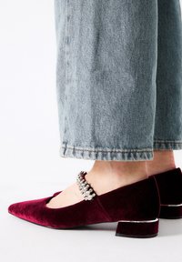 Tacones de terciopelo burdeos con punta afilada, adornados con una tira de cristal en el tobillo y un tacón bajo en bloque. Pantalones de mezclilla claros visibles.