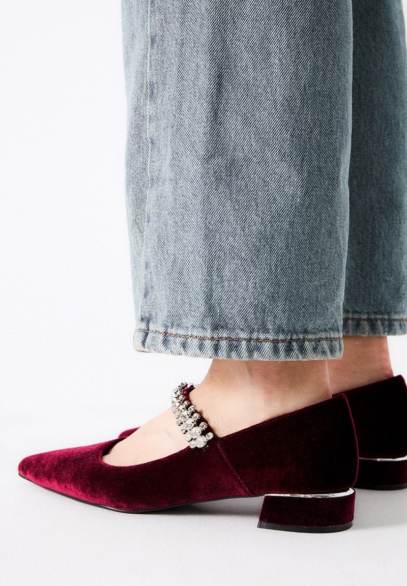 Tacones de terciopelo burdeos con punta afilada, adornados con una tira de cristal en el tobillo y un tacón bajo en bloque. Pantalones de mezclilla claros visibles.