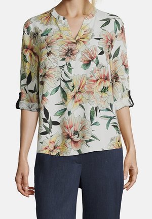 Femme portant une blouse blanche avec un grand imprimé floral multicolore et des manches retroussées, associée à un pantalon bleu foncé.