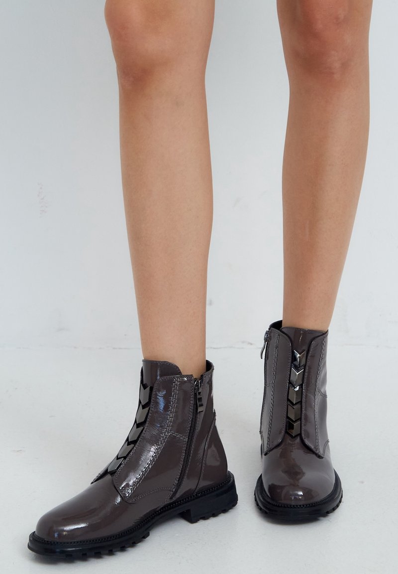 Stivaletti alla caviglia in pelle verniciata grigia con suola robusta e dettagli decorativi a cerniera sui lati. Texture liscia e design elegante.