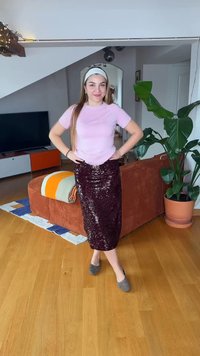 Jeune femme souriante, mains sur les hanches, portant un chemisier rose, une jupe à paillettes et un bandeau, dans un salon moderne avec des plantes et des meubles.