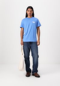 T-shirt en coton bleu clair à manches courtes avec un design imprimé sur la poitrine gauche, associé à un jean bleu et un tote bag de couleur cr�ème.