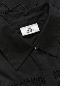 Sort skjorte med spidse kraver, detaljeret broderi og stofstruktur. En hvid etiket med et logo er synlig nær en knap.