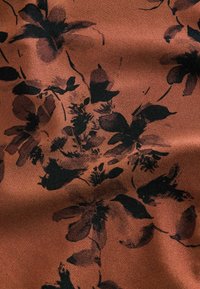 Tissu couleur rouille avec motif floral noir et brun foncé, présentant des fleurs et des feuilles abstraites avec une texture douce et de légers plis.