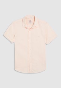 Camisa de manga corta con botones en suave tela color melocotón pastel. Presenta un cuello doblado y un logo sutil en el pecho izquierdo. Textura lisa.