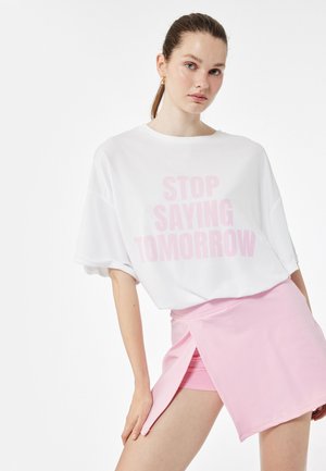 T-shirt bianca oversize con testo stampato in rosa "FERMATI DI DIRE DOMANI," abbinata a una mini gonna rosa chiaro con uno spacco laterale.