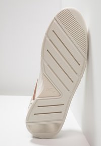 Suola di una scarpa da ginnastica, con una superficie in gomma beige testurizzata, caratterizzata da scanalature orizzontali e un accento con il logo, vista dal basso.
