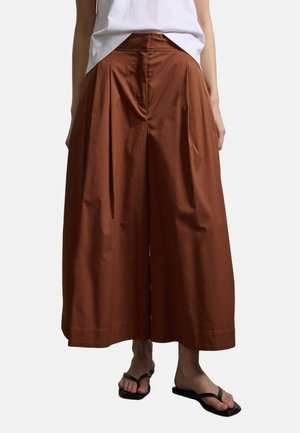 Frau trägt rostbraune, weit geschnittene Hosen mit Falten, ein weißes, hineingestecktes Oberteil und schwarze Flip-Flops vor weißem Hintergrund.