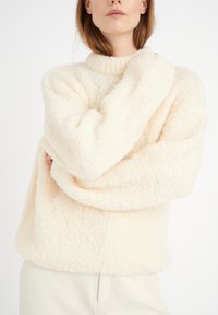 Pull crème duveteux avec un col montant côtelé et des manches larges. La texture est douce et moelleuse, offrant un aspect chaleureux.