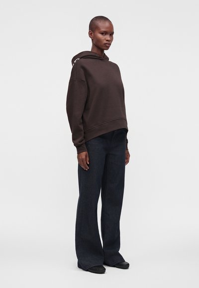 Personne debout, de face, portant un hoodie ample marron foncé, un jean large foncé et des chaussures noires sur un fond uni.