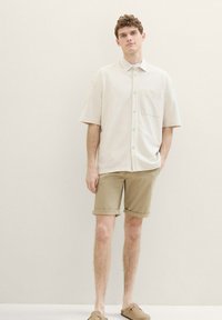 Cămașă bej cu mânecă scurtă și butoni, cu un buzunar în față, asortată cu pantaloni scurți kaki rulati și pantofi slip-on maro, pe un fundal neutru.