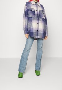Person in einer lila und weißen karierten, geknöpften Jacke, hellblauen Jeans und grünen Absatzschuhen vor einem weißen Hintergrund.