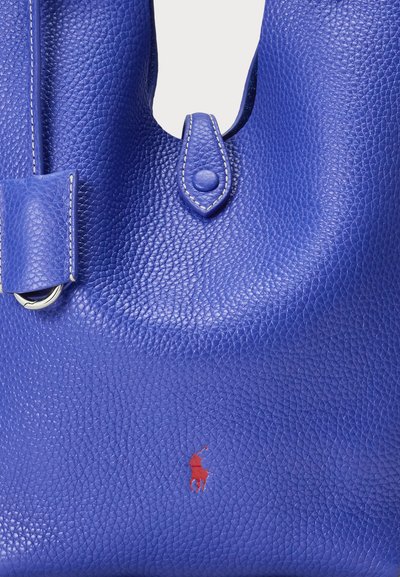 Zila ādas rokassoma ar strukturētu virsmu, kurai ir izteikta sarkana logo, augšējais aizdare un izturīgs roktura detaļa.