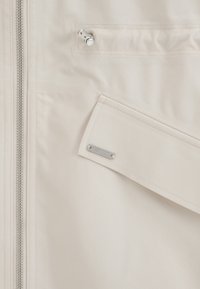 Détail de veste beige clair montrant une fermeture éclair argentée, une poche à rabat en biais avec une étiquette de marque en métal, et un cordon réglable avec loquet au-dessus de la poche.