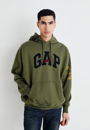 Giovane uomo che indossa un hoodie verde oliva della GAP con la scritta "Athletic", mani in tasca, in piedi davanti a uno sfondo bianco uniforme.