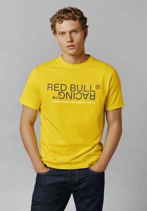 Żółta koszulka z napisem "RED BULL RACING" w pogrubionym czarnym kolorze. Krótkie rękawy, okrągły dekolt, materiał bawełniany, noszona z ciemnymi dżinsami.