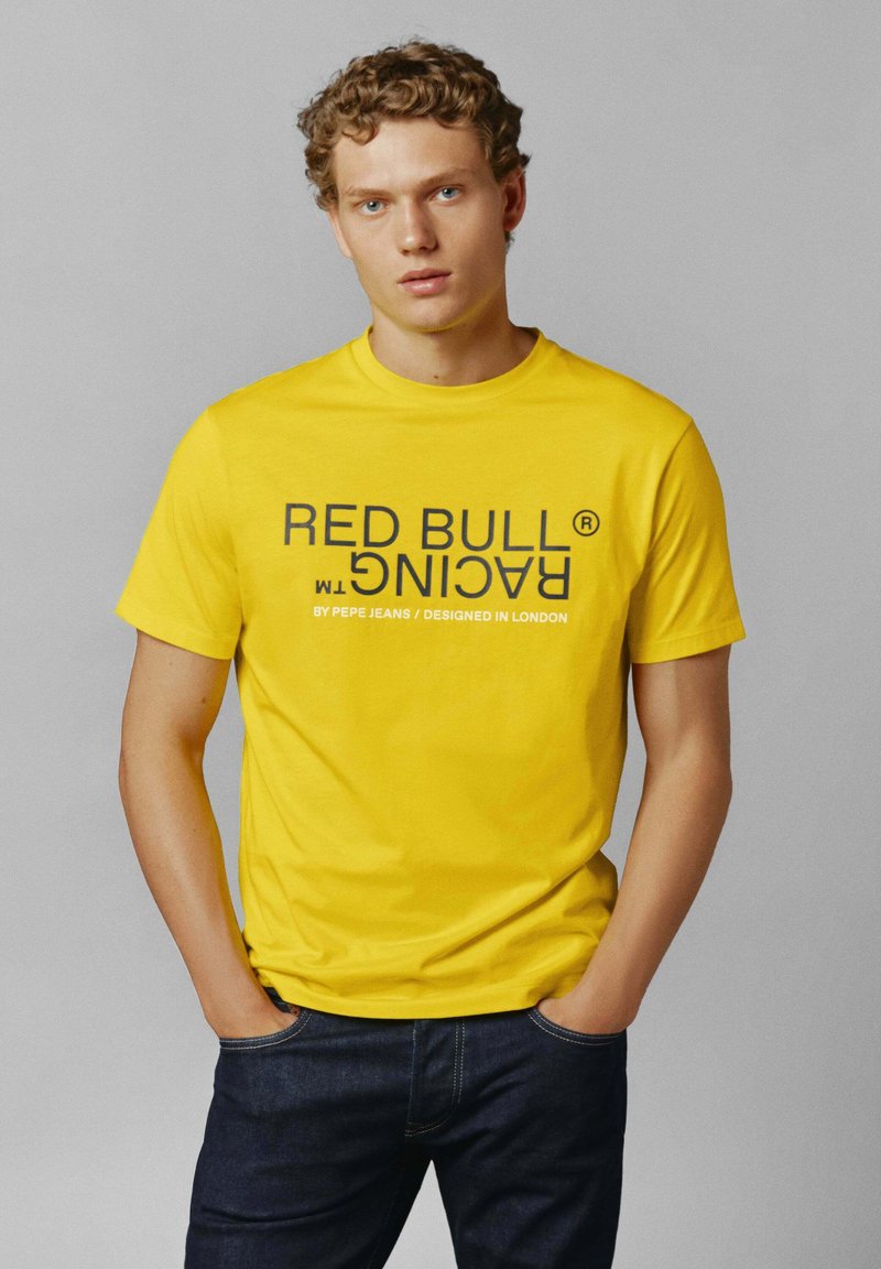 Žlté tričko s nápisom "RED BULL RACING" v tučnom čiernom písme. Krátke rukávy, okrúhly výstrih, materiál z bavlneného zmesi, nosené s tmavými džínsami.