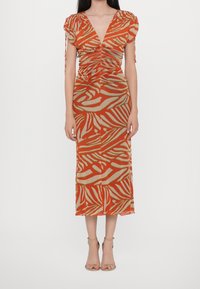 Femme portant une robe midi ajustée avec un imprimé zèbre abstrait orange et beige et des manches courtes froncées, associée à des escarpins nus à bride à la cheville.