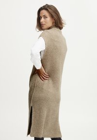 Beige gestrickte lange Weste mit einem strukturierten Muster, seitlichen Schlitzen, ärmellosem Design und hohem Kragen, getragen über einem weißen Oberteil.