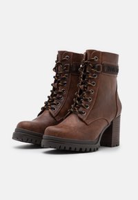 Stivali alla caviglia in pelle marrone con robusti tacchi impilati, lacci neri e una cinghia decorativa etichettata "TOM TAILOR." Finitura testurizzata e suole rigate.