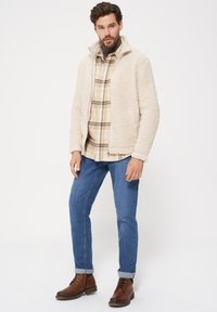 Beżowa polarowa kurtka z wysokim kołnierzem, noszona na koszuli w kratę. Model połączył ją z niebieskimi dżinsami i brązowymi, sznurowanymi botkami. Prosty, casualowy design.